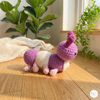 A handmade crocheted caterpillar toy in purple, cream, and pink, placed on a wooden floor in a sunlit room with potted plants in the background.
