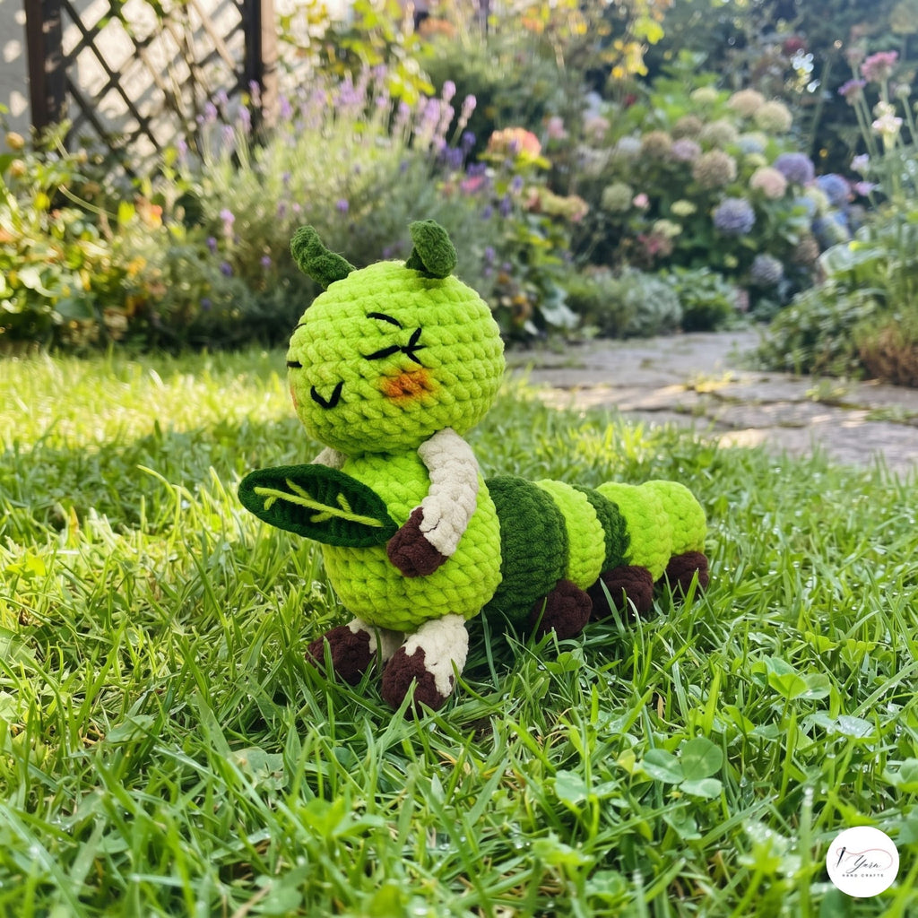 A handmade crocheted caterpillar toy sitting on a green lawn in a garden setting. The toy features a bright green body, dark green stripes, cream-colored arms and legs, and a small orange nose.