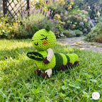 A handmade crocheted caterpillar toy sitting on a green lawn in a garden setting. The toy features a bright green body, dark green stripes, cream-colored arms and legs, and a small orange nose.