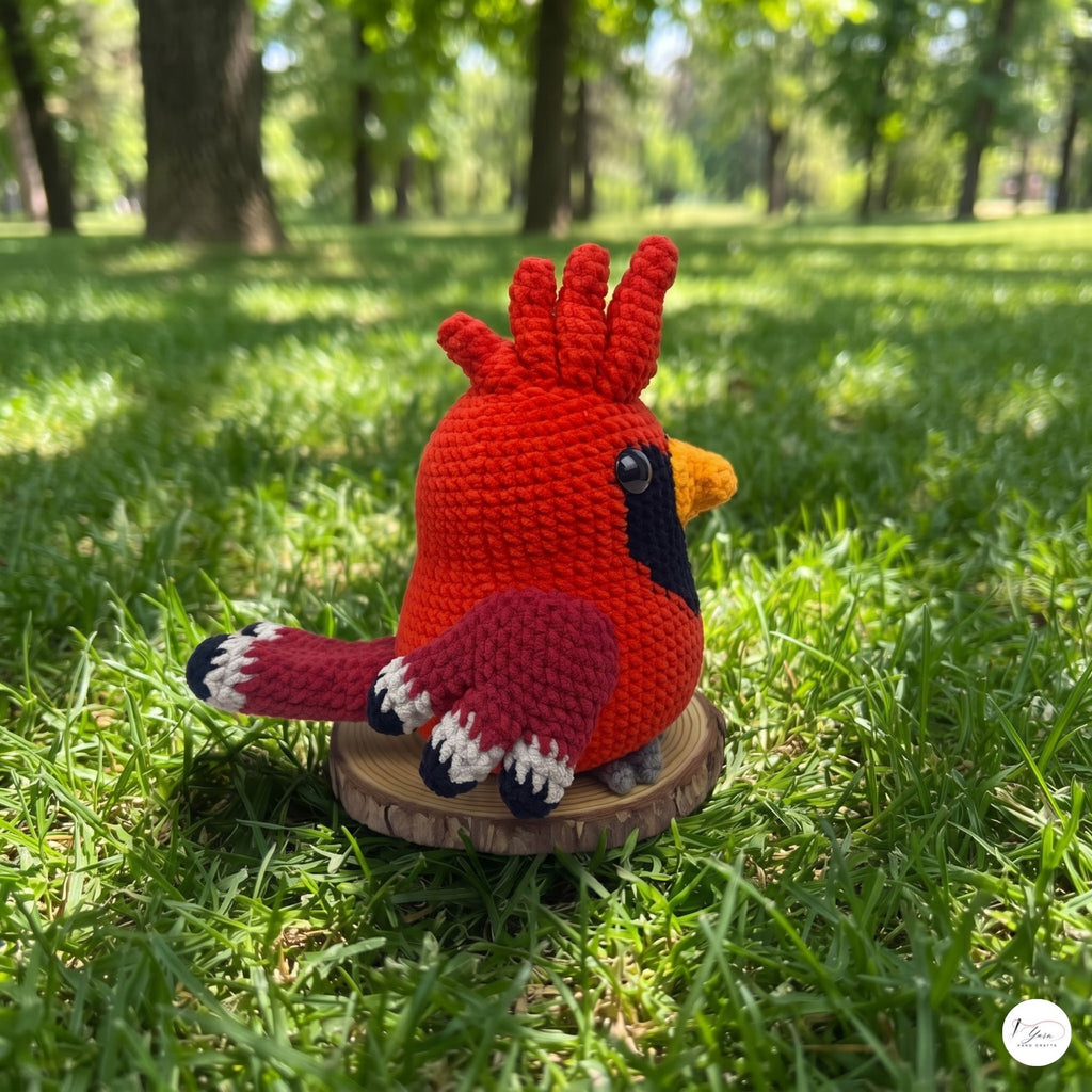 A crocheted cardinal bird toy in bright red, with a black face, yellow beak, and white-tipped tail feathers, sitting on a small round wooden slice in a grassy park setting.