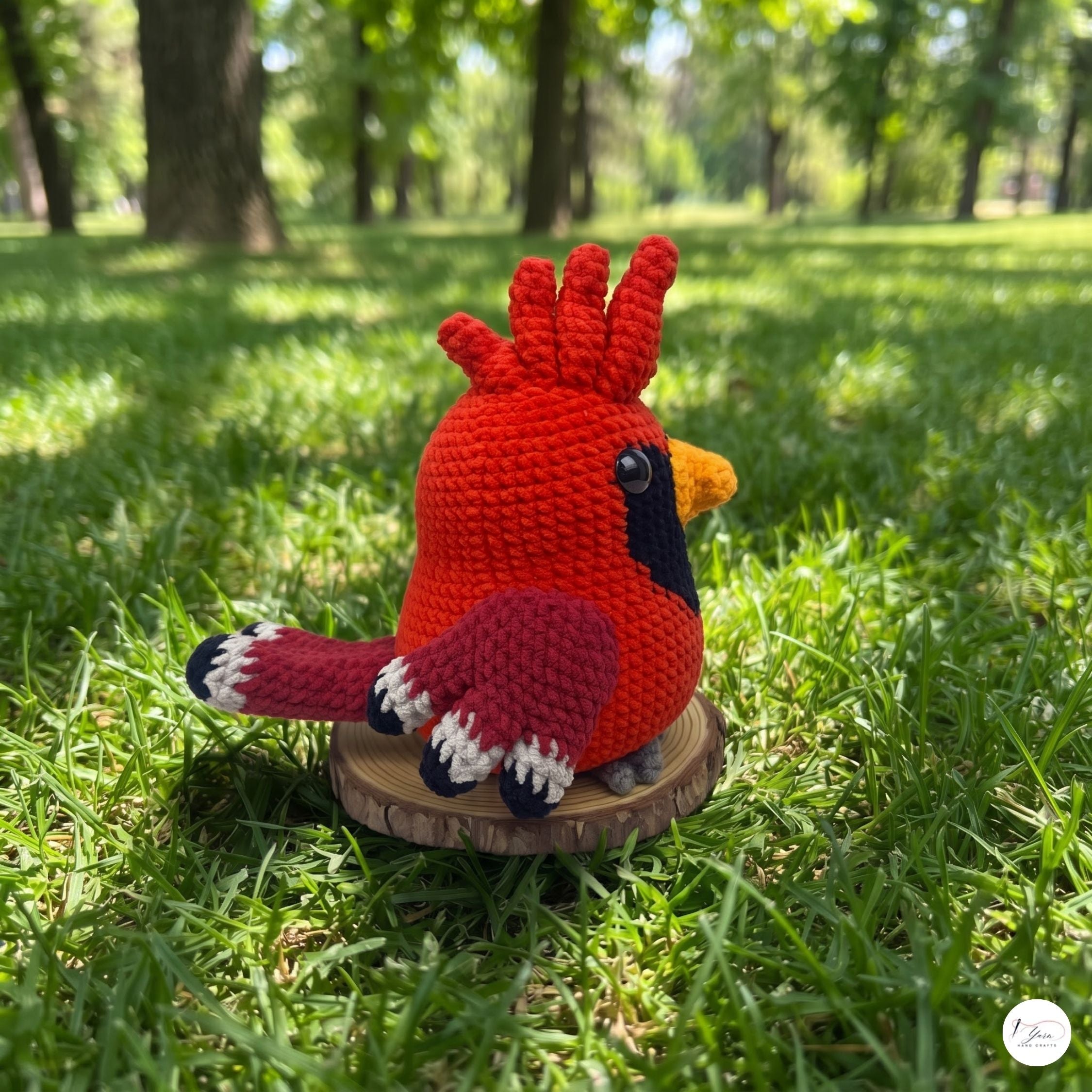 A crocheted cardinal bird toy in bright red, with a black face, yellow beak, and white-tipped tail feathers, sitting on a small round wooden slice in a grassy park setting.