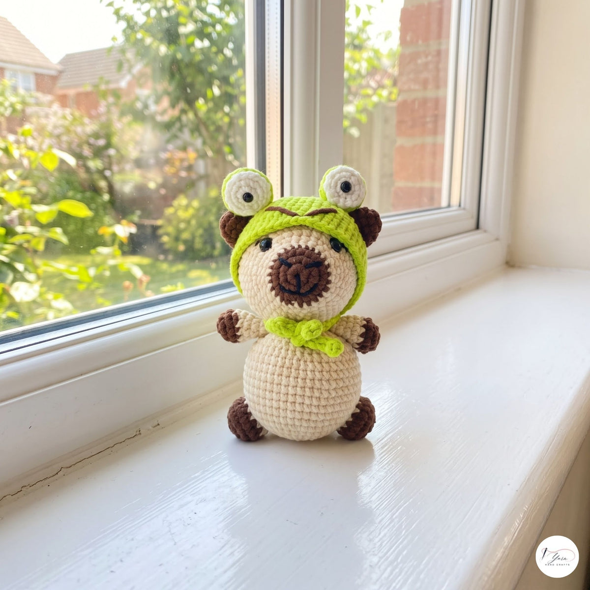 A crocheted bear toy wearing a green frog hat and bow tie, sitting on a white windowsill with a garden view in the background.