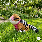 A crocheted tiger doll with orange, black, and white stripes, a pink scarf, and a pink nose sits on green grass in a garden setting.