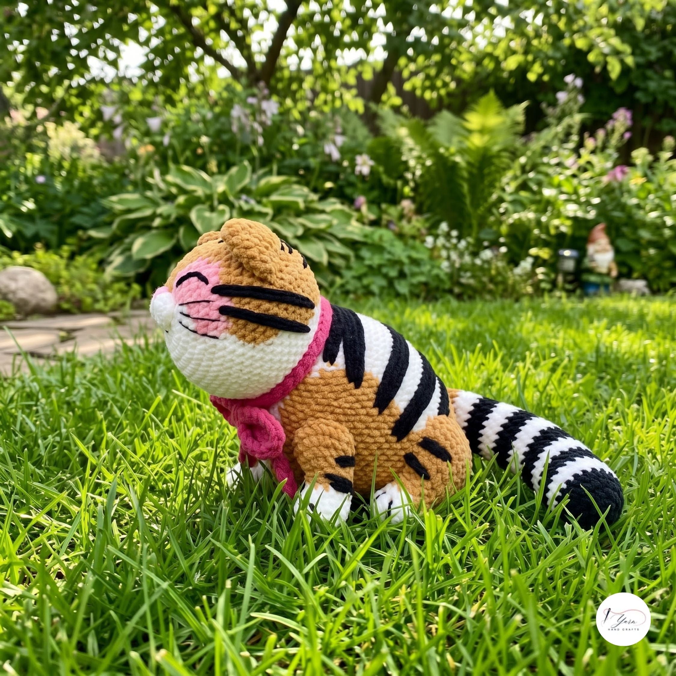 A crocheted tiger doll with orange, black, and white stripes, a pink scarf, and a pink nose sits on green grass in a garden setting.