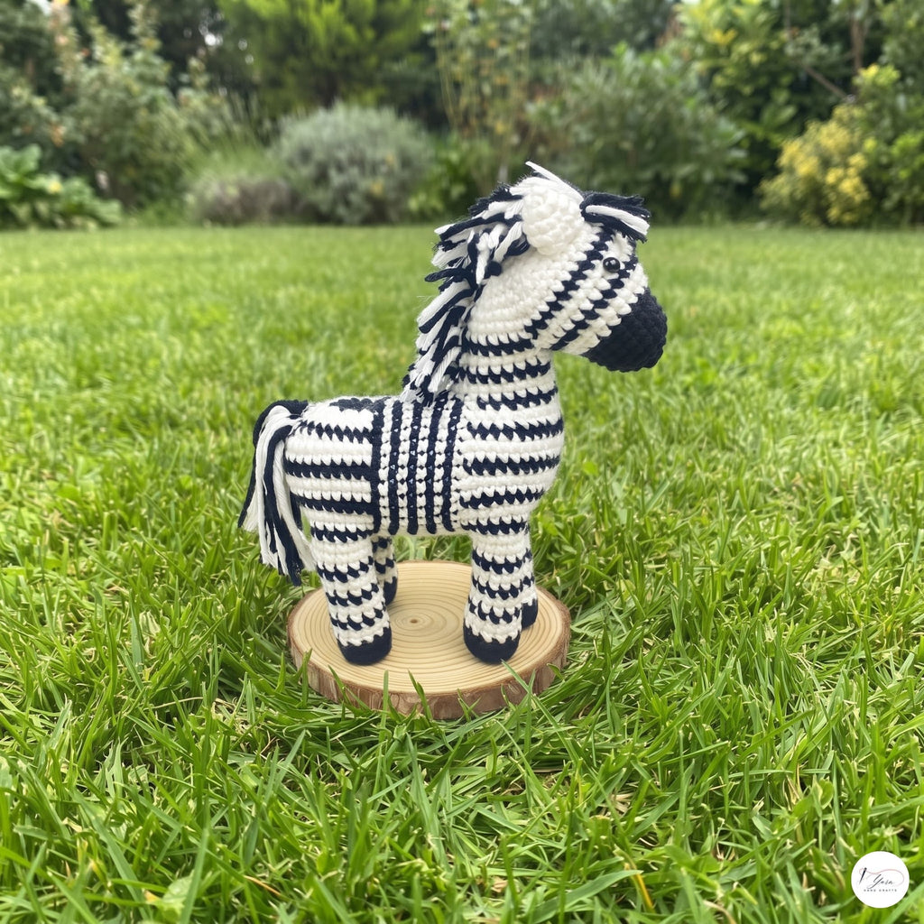 A crocheted zebra toy standing on a small round wooden base in a grassy outdoor garden setting.