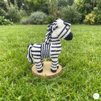 A crocheted zebra toy standing on a small round wooden base in a grassy outdoor garden setting.