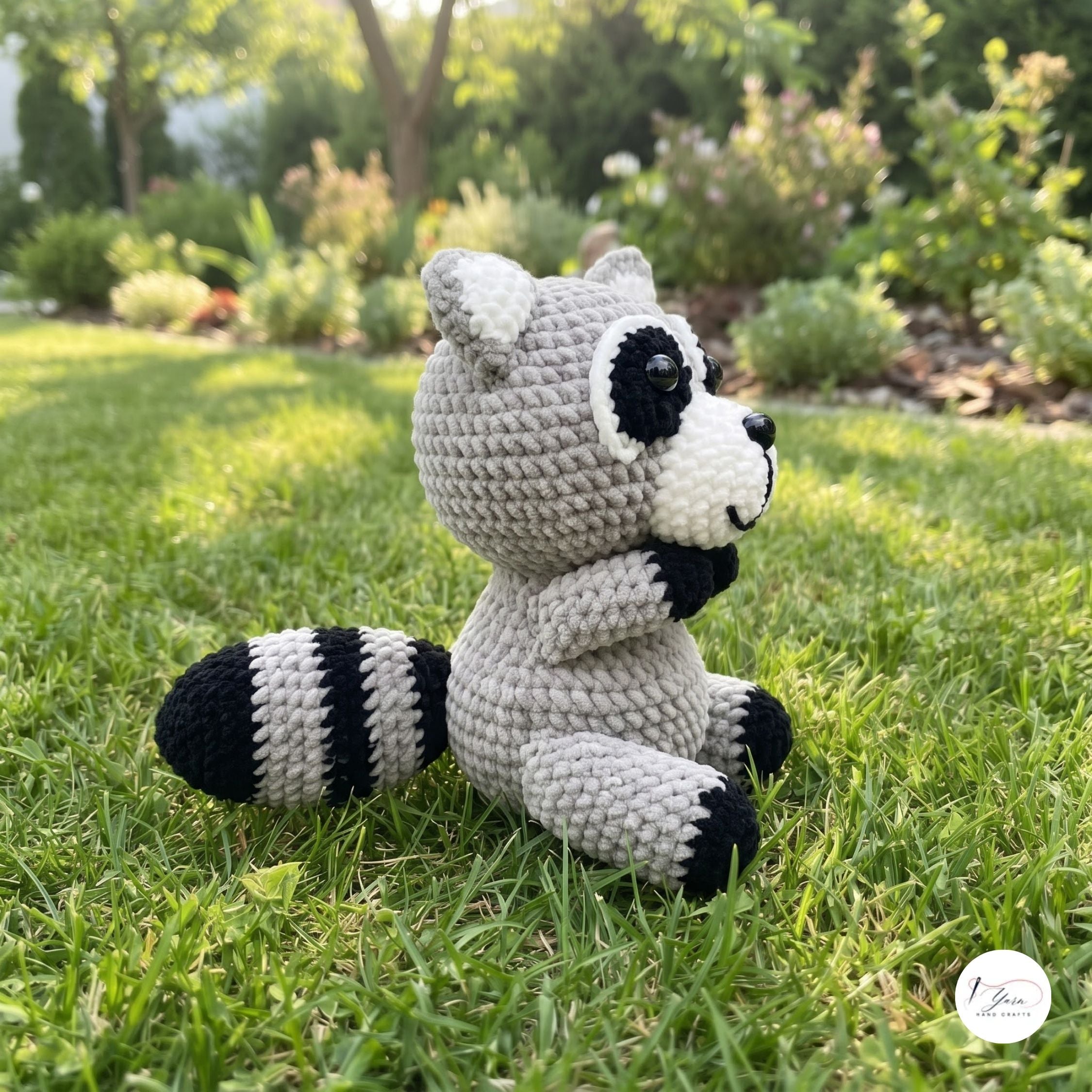 A crocheted raccoon plush toy sitting on a green grass lawn in a sunny garden. The toy is made of light gray yarn with black and white accents on its face, paws, and striped tail.