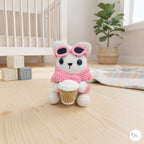 A cute handmade white crochet cat plush sitting on a wooden floor in a cozy nursery setting, wearing a pink sweater, pink sunglasses pushed up on its head, and holding a crocheted takeaway coffee cup.