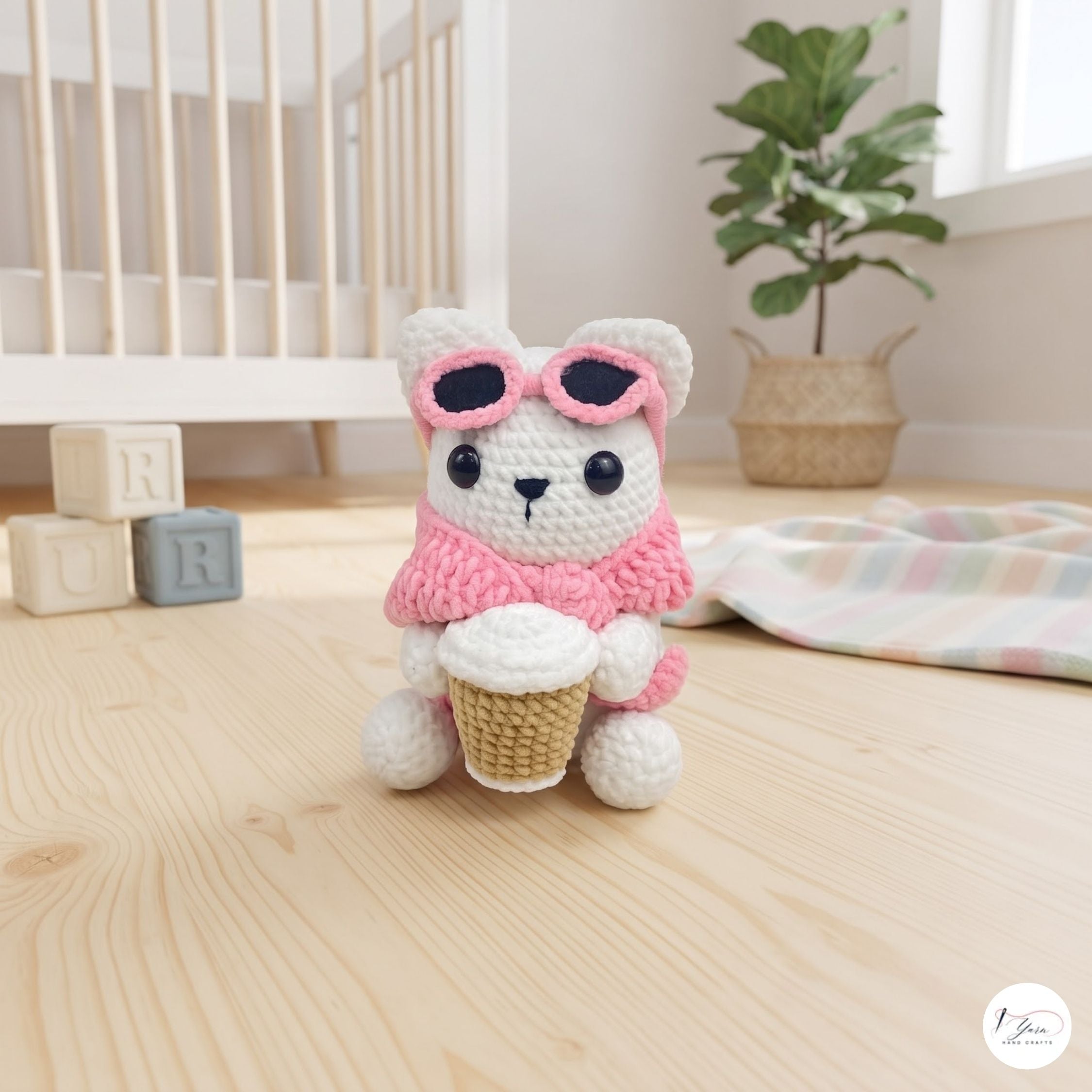A cute handmade white crochet cat plush sitting on a wooden floor in a cozy nursery setting, wearing a pink sweater, pink sunglasses pushed up on its head, and holding a crocheted takeaway coffee cup.