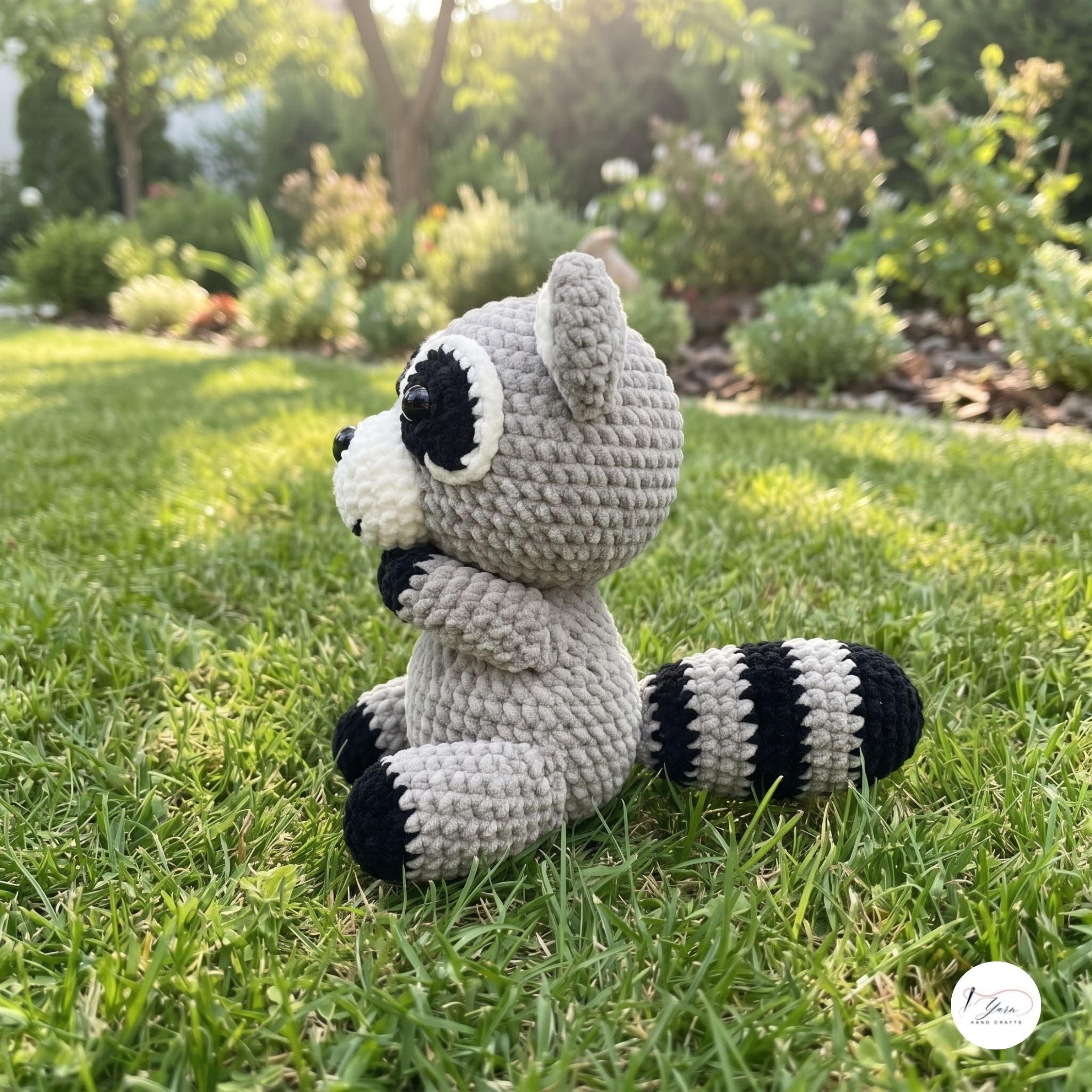 A crocheted raccoon plush toy sitting on a green grass lawn in a sunny garden setting. The toy is made of light gray yarn with black and white accents on its face, paws, and striped tail.