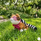 A crocheted tiger doll with orange, black, and white stripes sits on green grass in a garden setting.