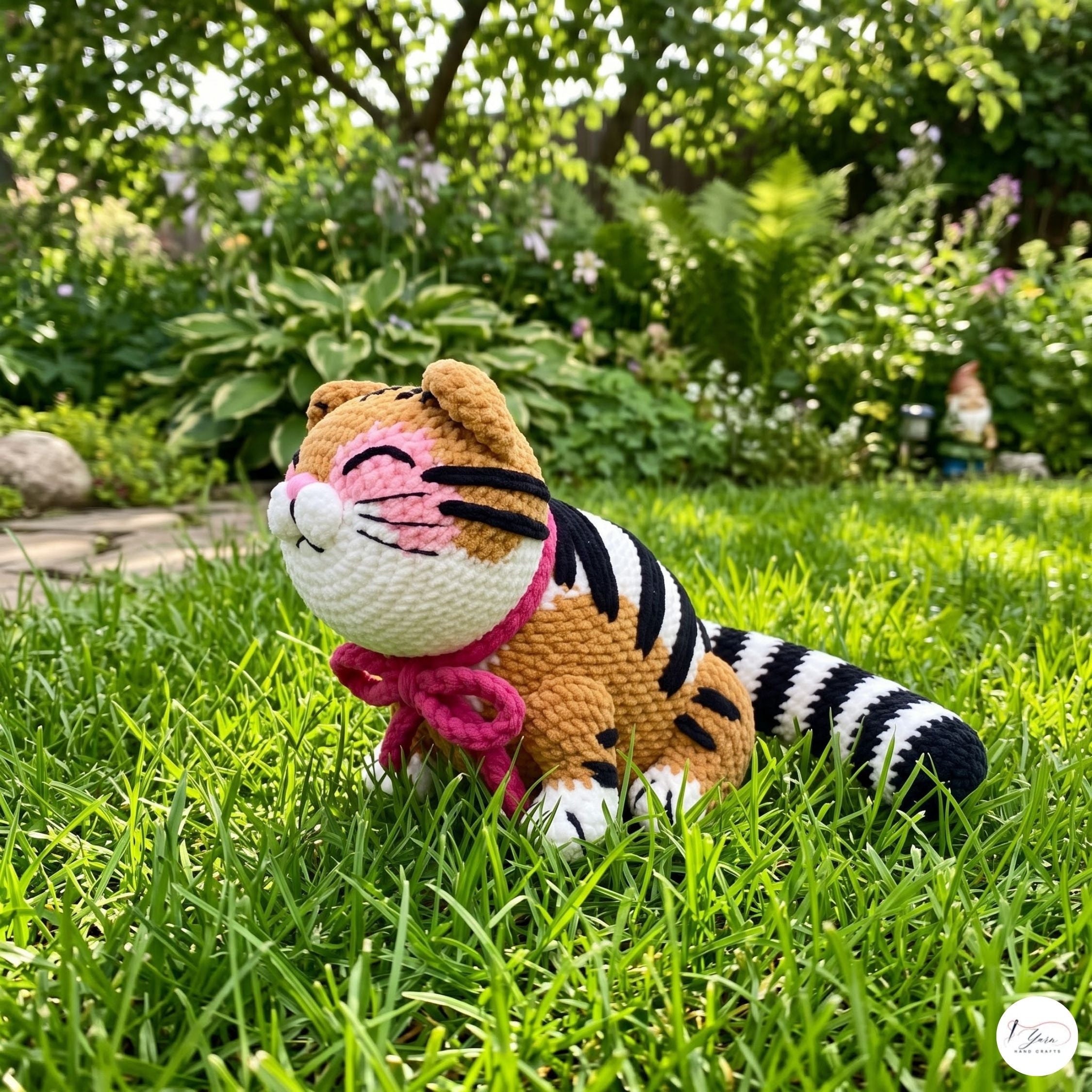 A crocheted tiger doll with orange, black, and white stripes sits on green grass in a garden setting.