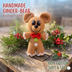 A handmade crocheted ginger bear toy stands on a rustic wooden surface surrounded by pine branches, holly leaves, and red berries. Text in the upper left reads “HANDMADE GINGER-BEAR Festive Friend,” and text in the lower right reads “100% COTTON SOFT & SAFE.