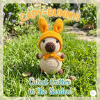 A crocheted bunny doll wearing a yellow cap and matching scarf, standing on green grass outdoors with flowers and a garden path in the background. Text overlays read 'CAPY-BUNNY!' and 'Cutest Critter in the Garden!'.