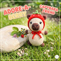 A crocheted capybara doll wearing a red strawberry-themed hood sits on grass beside a stone with small flowers and berries. Text overlays read “ADORE-A BERRY” and “CAPYBARA FRIEND.