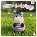 A crocheted plush donkey toy labeled 'CUDDLY Crochet Donkey' stands on green grass outdoors. The donkey is lavender-gray with a cream face and neck, black nose and hooves, and small dark ears.