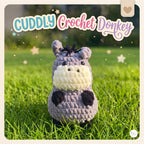 A crocheted plush donkey toy labeled 'CUDDLY Crochet Donkey' stands on green grass outdoors. The donkey is lavender-gray with a cream face and neck, black nose and hooves, and small dark ears.
