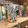 A handmade crocheted zebra baby doll with black and white chevron stripes, standing on a round wooden slice base on a wooden shelf. Books and small wooden animal toys are blurred in the background.