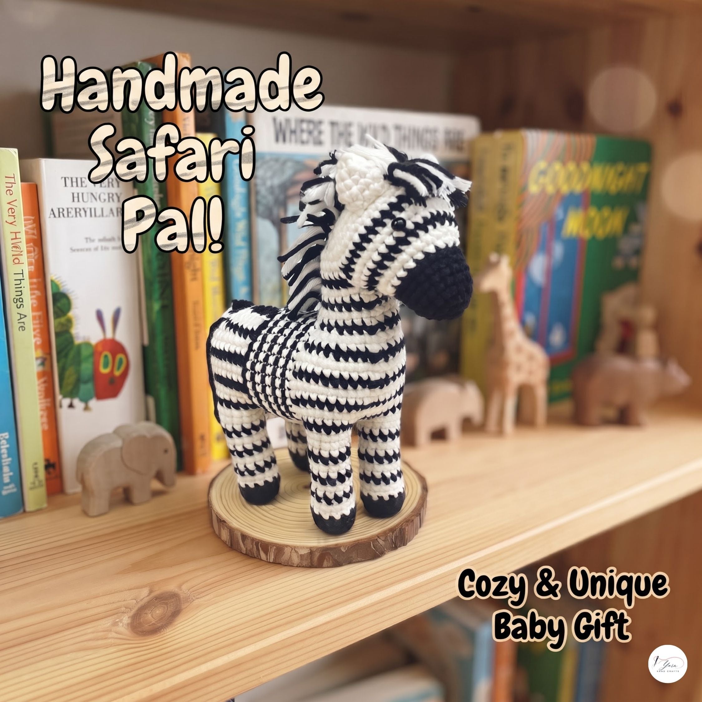 A handmade crocheted zebra baby doll with black and white chevron stripes, standing on a round wooden slice base on a wooden shelf. Books and small wooden animal toys are blurred in the background.