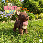 A crocheted puppy doll stands on green grass in a sunny garden, holding a pink heart. Large overlaid text reads 'MY HEART PUPPY Handmade Cuddles.