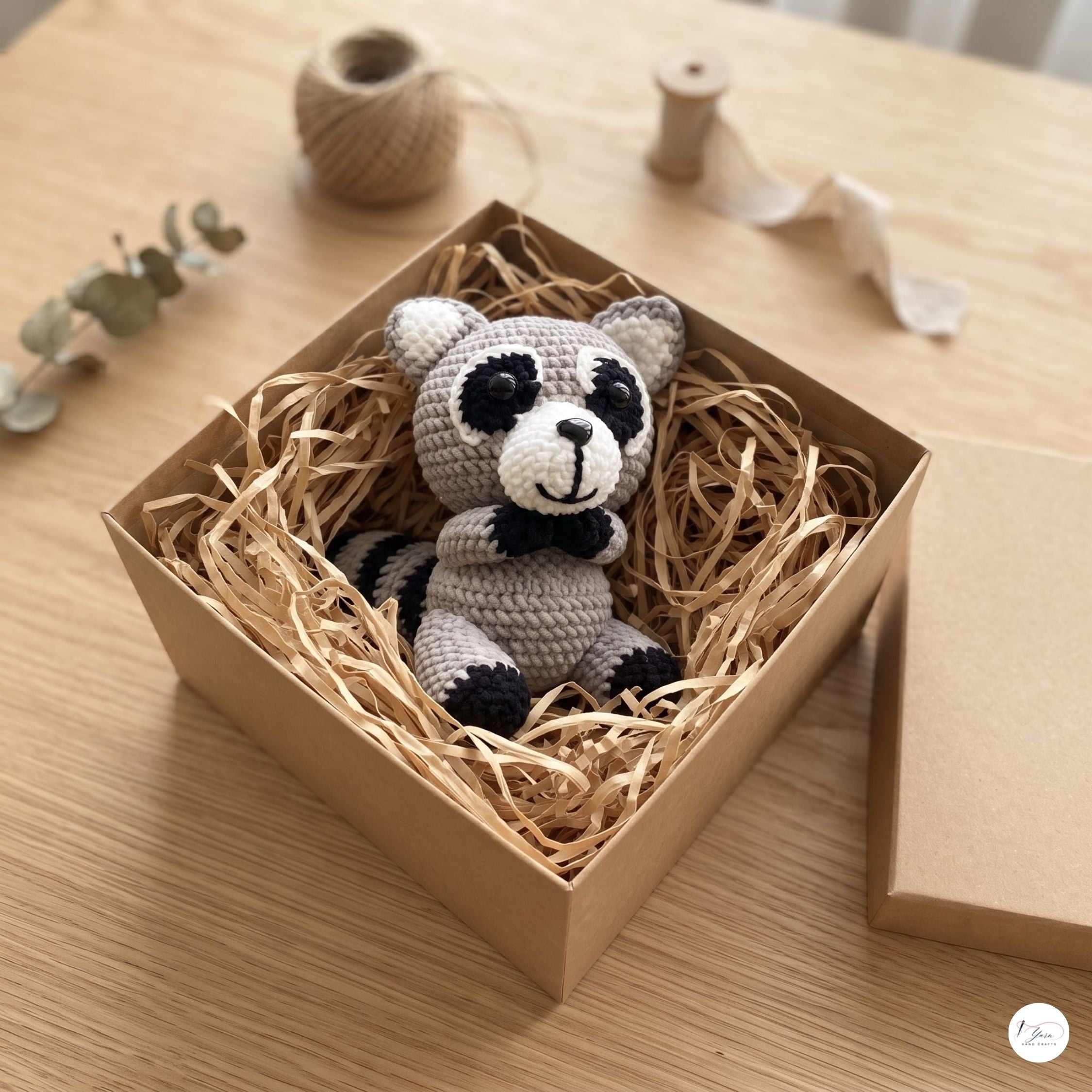 A crocheted raccoon toy sits inside a kraft paper gift box filled with shredded paper on a wooden table. The raccoon is light gray with black and white facial features and black-and-white striped tail patches.