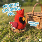 A crocheted cardinal bird figure in bright red with a black chest, yellow beak, and pink wing, displayed on a small wooden slice outdoors on grass next to a wicker basket. Large text reads 'CROCHET CARDINAL PAL' and 'HANDMADE FRIEND'.