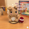 A cute crocheted plush bunny toy made from tan yarn sits on a wooden slice base on a wooden desk, next to a small red-and-white crocheted egg. The background shows a softly blurred room with books and colored pencils, and the text 'Meet Your New CROCHET BUDDY!' appears above.