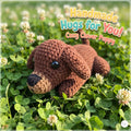 A handmade crocheted puppy toy lying on green clover with white flowers. The text overlay reads 'Handmade Hugs for You! Cozy Clover Puppy'.