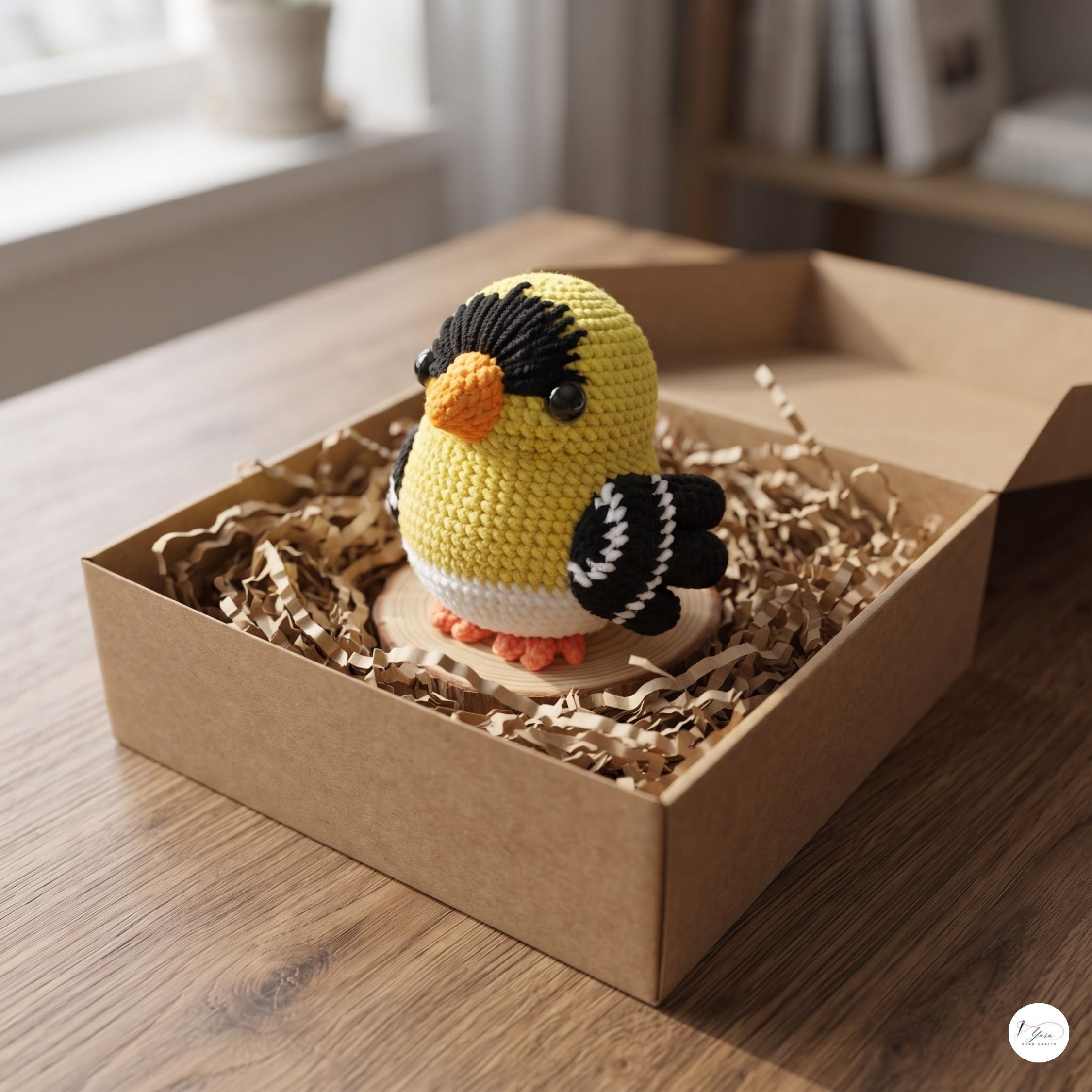 A crocheted bird toy in yellow, black, white, and orange sits inside a brown cardboard box filled with shredded paper on a wooden table.
