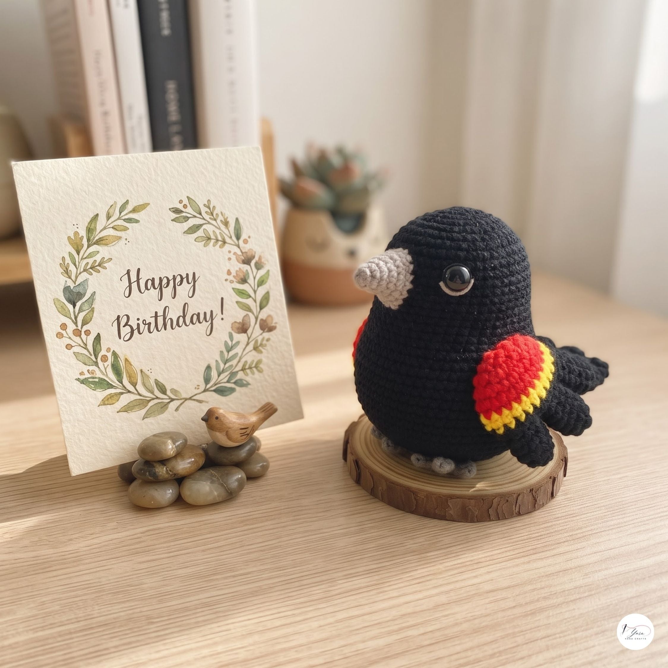 A crocheted black bird figurine with red and yellow wing accents stands on a small wooden slice base on a tabletop. Beside it is a greeting card that reads “Happy Birthday!” with a floral wreath design, styled with small stones and a potted succulent in the background.