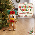 A crocheted gingerbread duck doll stands on a wooden floor beside a lit Christmas tree. A decorative sign reads “Gingerbread Duck The Cutest Christmas Creation.
