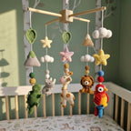 A crocheted baby mobile with wooden hanging bars, featuring colorful handmade plush toys including stars, mountains, a crocodile, giraffe, lion, parrot, and clouds, displayed in a nursery crib against a green wall.
