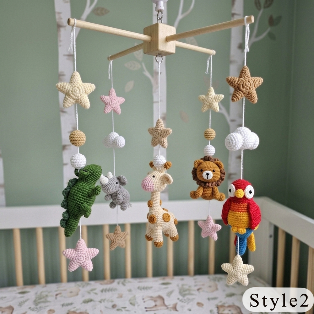 A crocheted baby mobile hanging in a crib, featuring animal and nature shapes including a green crocodile, giraffe, lion, parrot, stars, clouds, and wooden star accents on a wooden crossbar. The crib is set against a soft green wall with white birch tree decals.