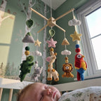 A baby sleep crib with a sleeping infant in the foreground and a crocheted hanging mobile above. The mobile features a light wood star frame with knitted animals and clouds in green, pink, yellow, brown, red, blue, and white, set against a green wall with birch tree wallpaper.