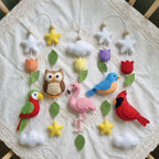 A handmade felt baby nursery decoration set featuring colorful animal and nature shapes including a red cardinal, pink flamingo, green and red parrot, brown owl, blue bird, white clouds, yellow and purple flowers, and green leaves, all arranged on a cream lace-trimmed fabric background.