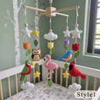 A wooden baby crib mobile with felt birds, flowers, stars, and clouds hanging in a nursery. The mobile features a pink flamingo, a red cardinal, a green and red parrot, a brown owl, a blue bird, white stars, white and white cloud shapes, green and yellow flowers, and green leaves, suspended from a light wood crossbar over a white crib.