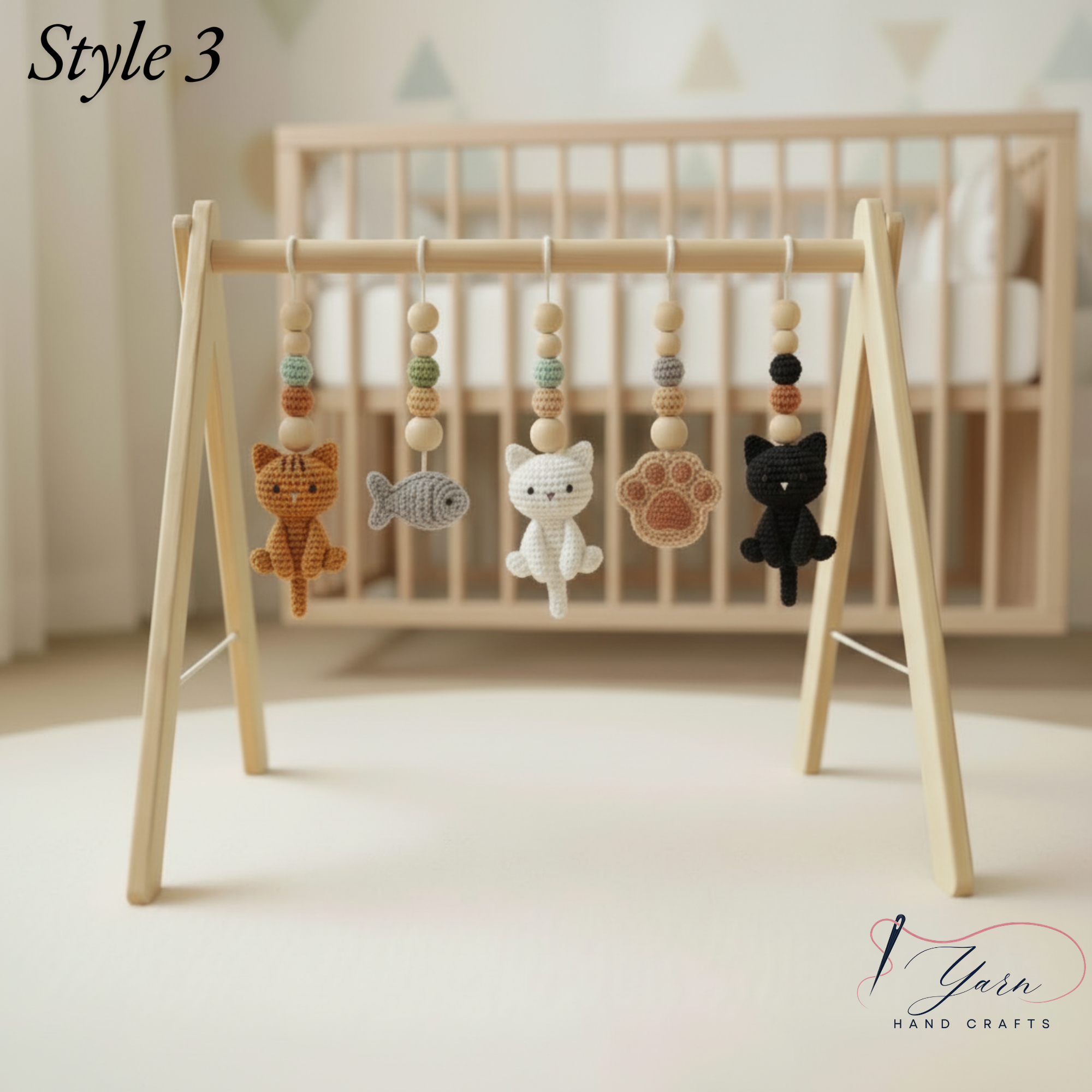 A wooden A-frame baby toy stand with hanging yarn crafts shaped like a cat, fish, white cat, paw print, and black cat, shown in a softly lit nursery setting with a crib in the background. The text 'Style 3' appears in the top left, and a 'Yarn Hand Crafts' logo is in the bottom right.