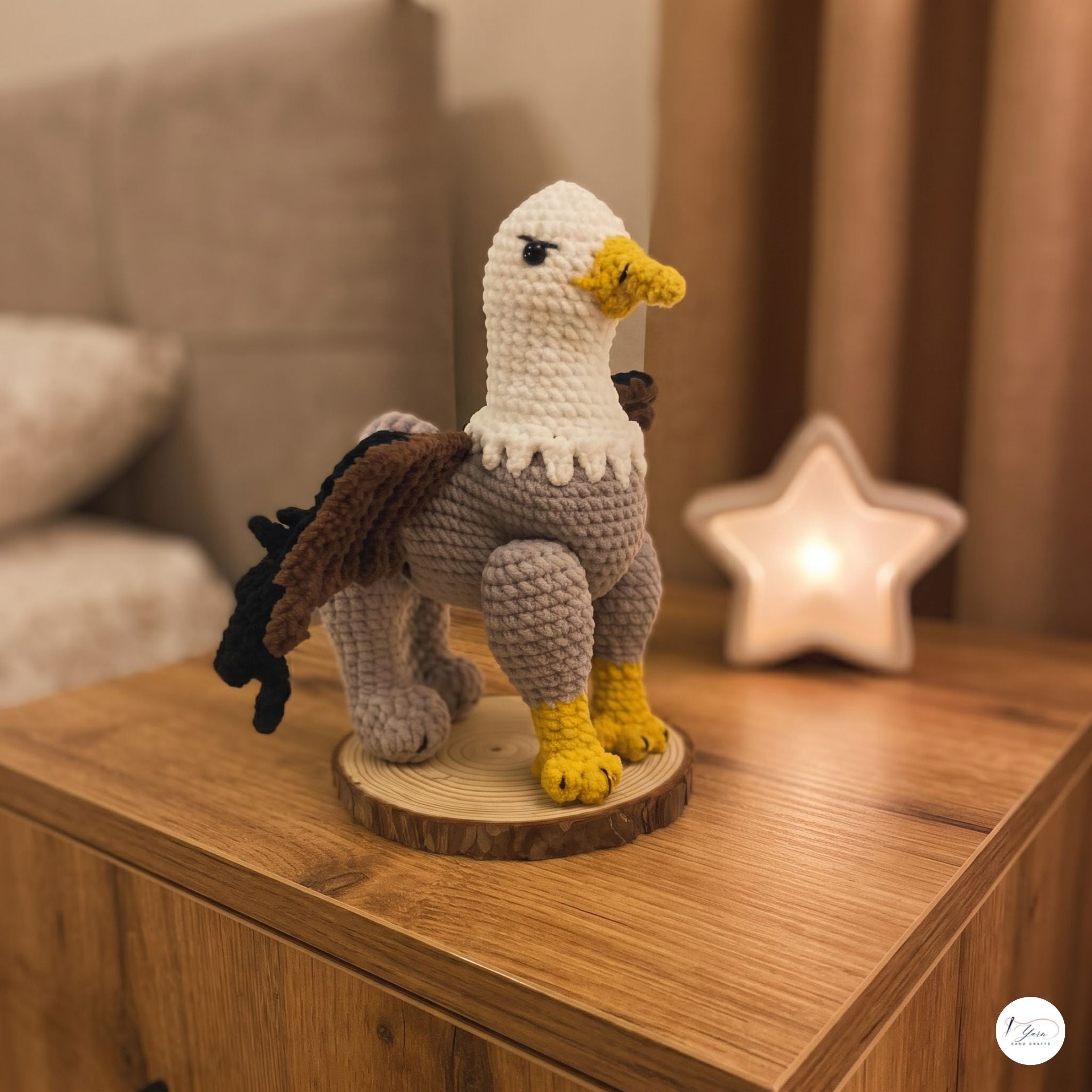A handmade crocheted eagle figurine standing on a round wooden slice coaster, placed on a wooden bedside table next to a glowing star-shaped night light.