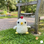 A small crocheted chick toy sits on green grass beside a weathered wooden bench in a blurred outdoor garden setting.