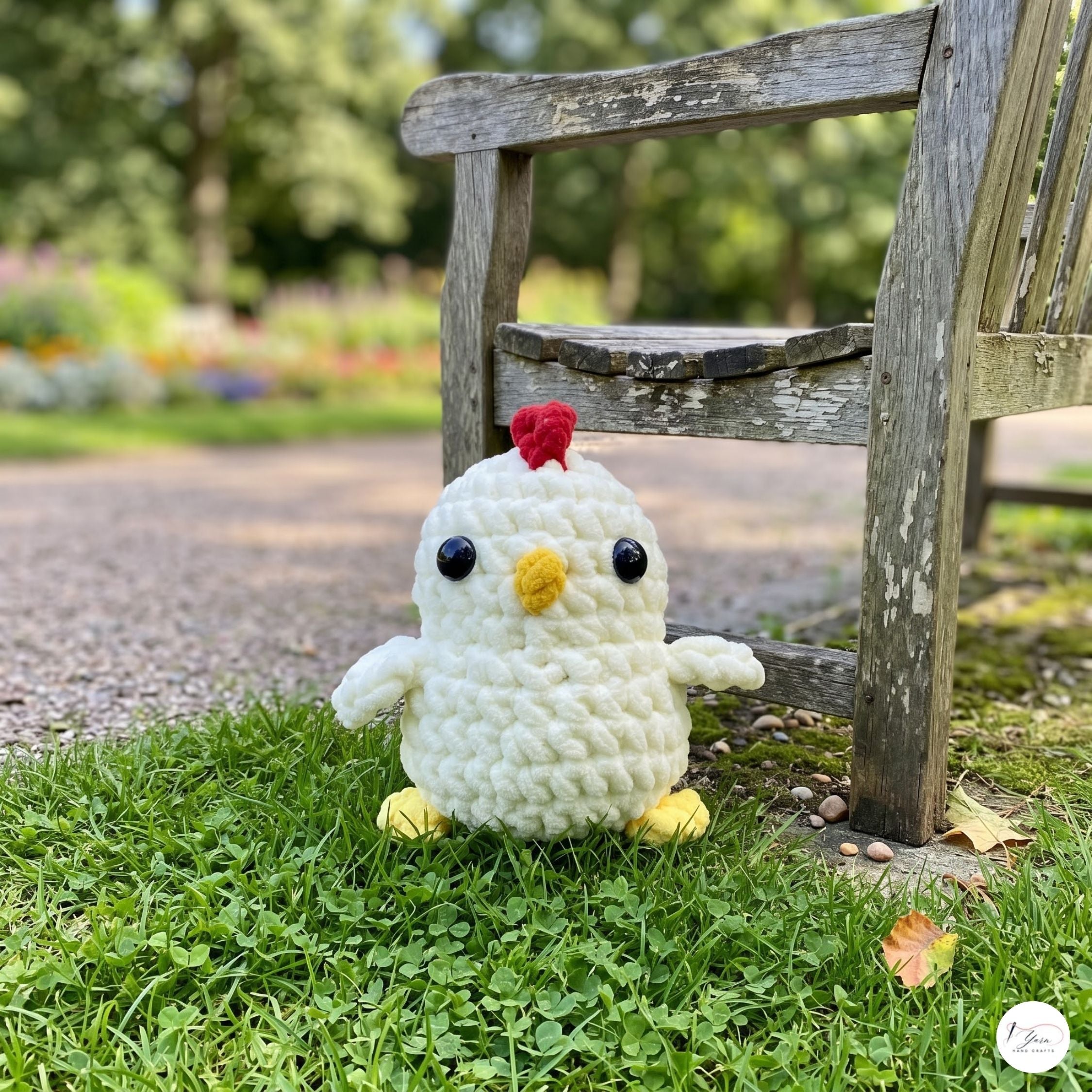 A small crocheted chick toy sits on green grass beside a weathered wooden bench in a blurred outdoor garden setting.