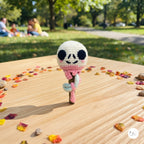 A crocheted amigurumi-style character figure with a white head, black eyes, pink scarf, and brown stick-like body stands on a wooden table outdoors. The background shows a blurred park scene with green grass, trees, and people sitting in the distance.