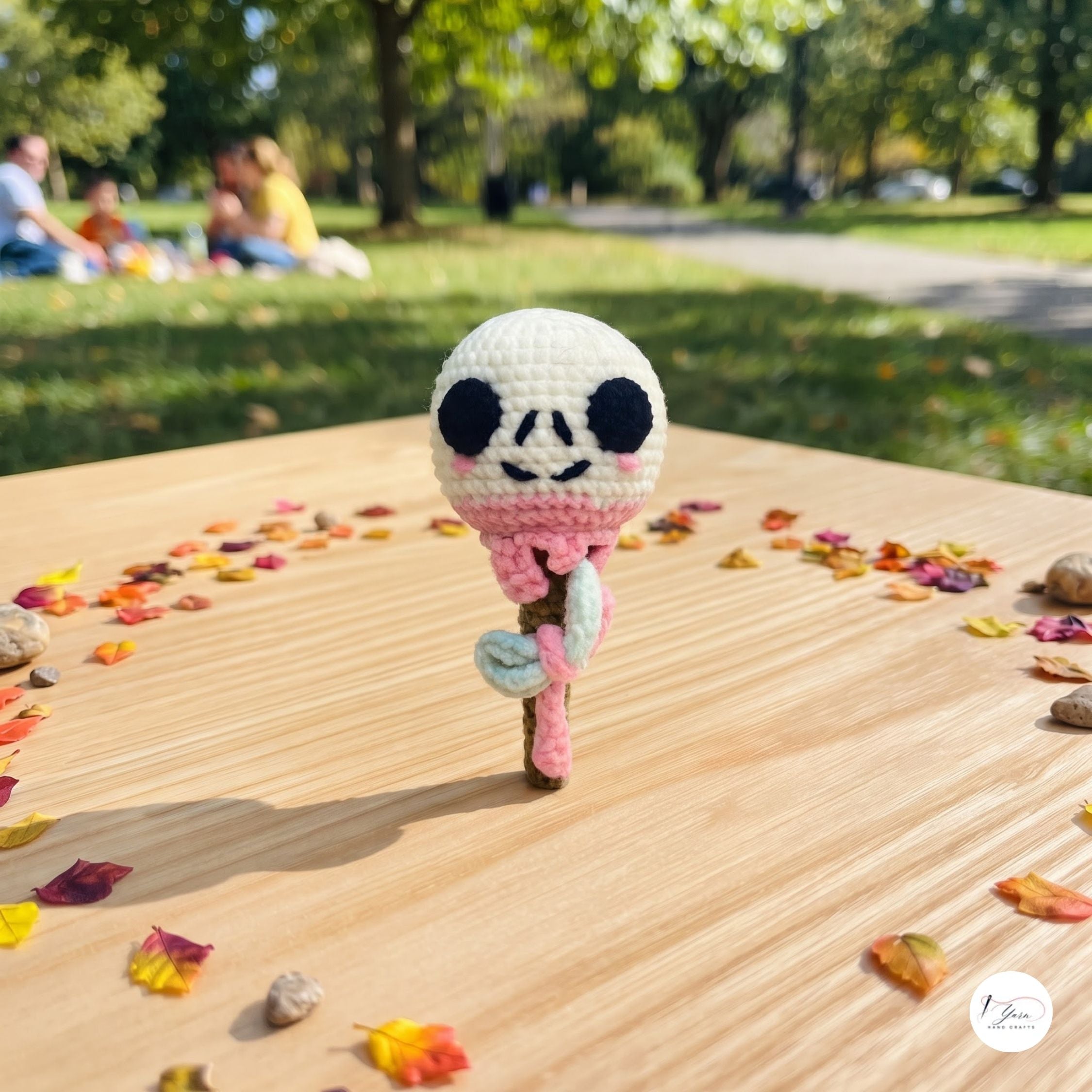 A crocheted amigurumi-style character figure with a white head, black eyes, pink scarf, and brown stick-like body stands on a wooden table outdoors. The background shows a blurred park scene with green grass, trees, and people sitting in the distance.