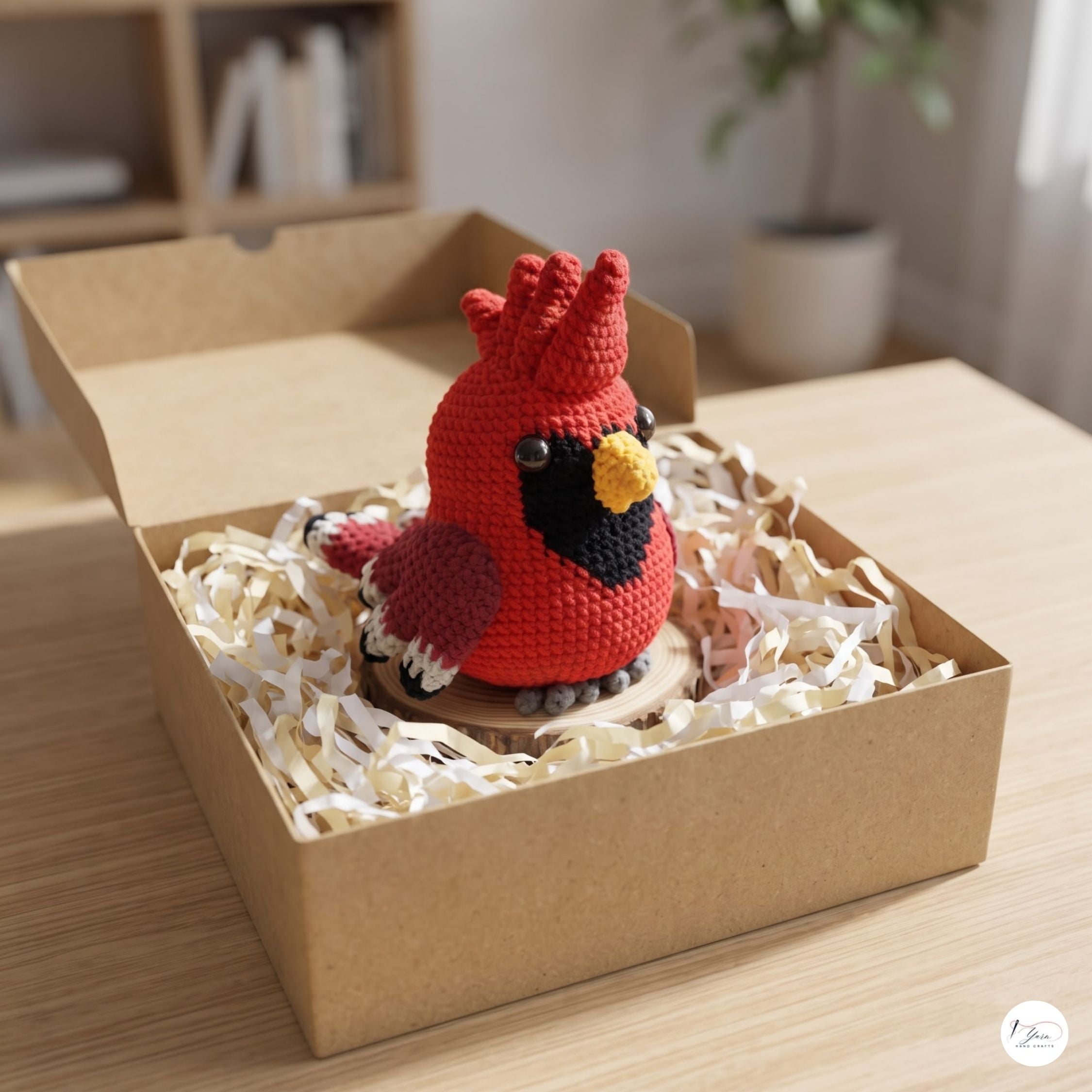 A crocheted red cardinal bird ornament displayed inside an open brown cardboard box filled with white paper shred, set on a light wood tabletop in a softly blurred indoor setting.