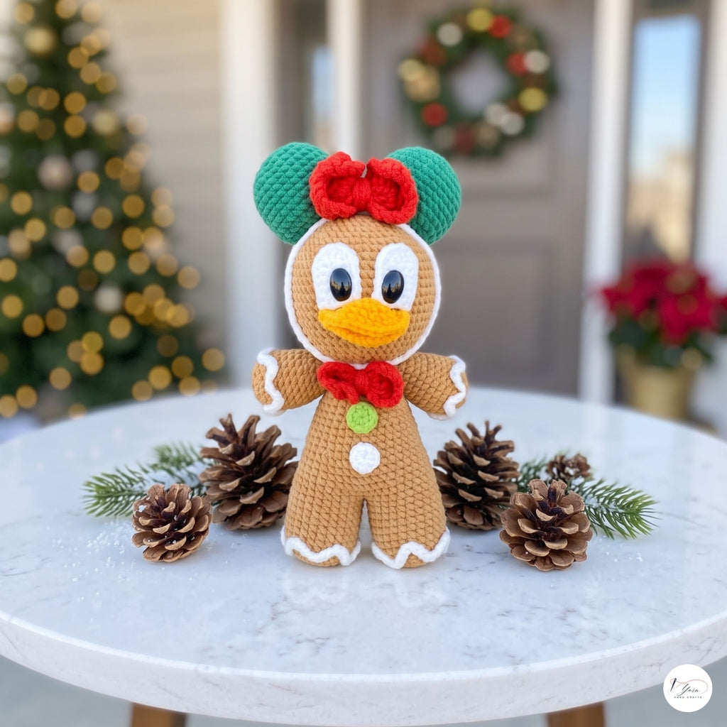 A crocheted gingerbread character decoration sits on a round white table, surrounded by pinecones and evergreen branches. The character has green ears, a red bow, a red bow tie, and a white button, with a blurred Christmas tree and wreath in the background.