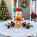 A crocheted gingerbread character decoration sits on a round white table, surrounded by pinecones and evergreen branches. The character has green ears, a red bow, a red bow tie, and a white button, with a blurred Christmas tree and wreath in the background.