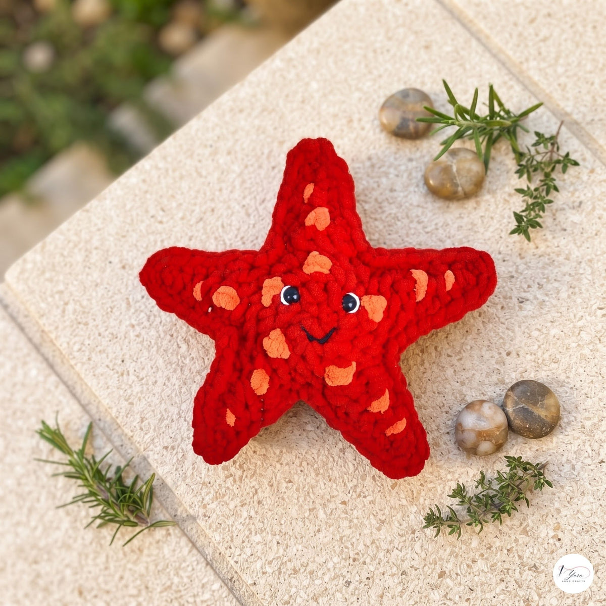 A red crocheted starfish with a smiling face and orange spots, styled on a beige stone surface with small green sprigs and stones.