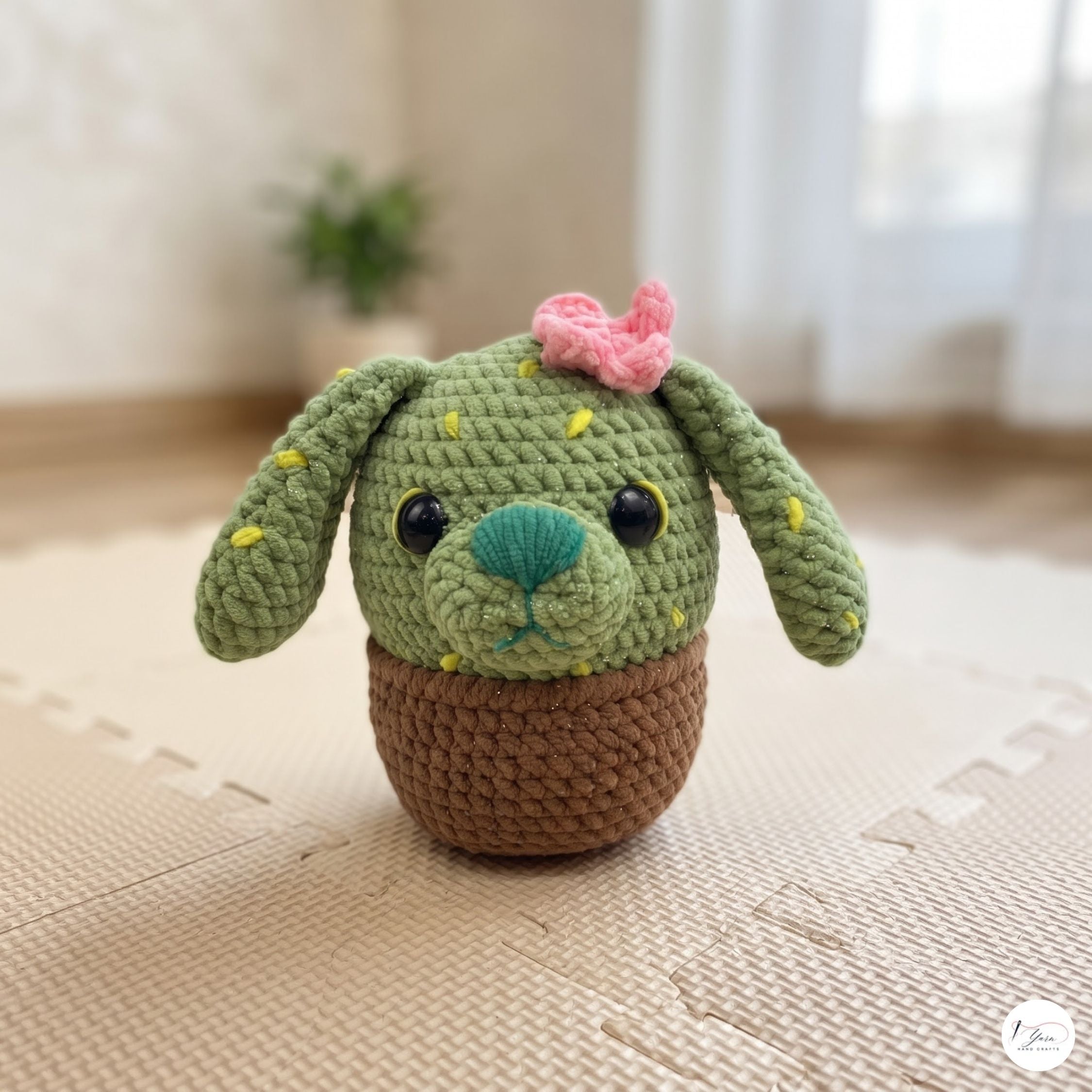 A crocheted plush toy shaped like a cactus dog, featuring a green textured body with yellow cactus details, floppy ears, a pink bow, and a brown base, set on a beige play mat in a softly blurred indoor background.