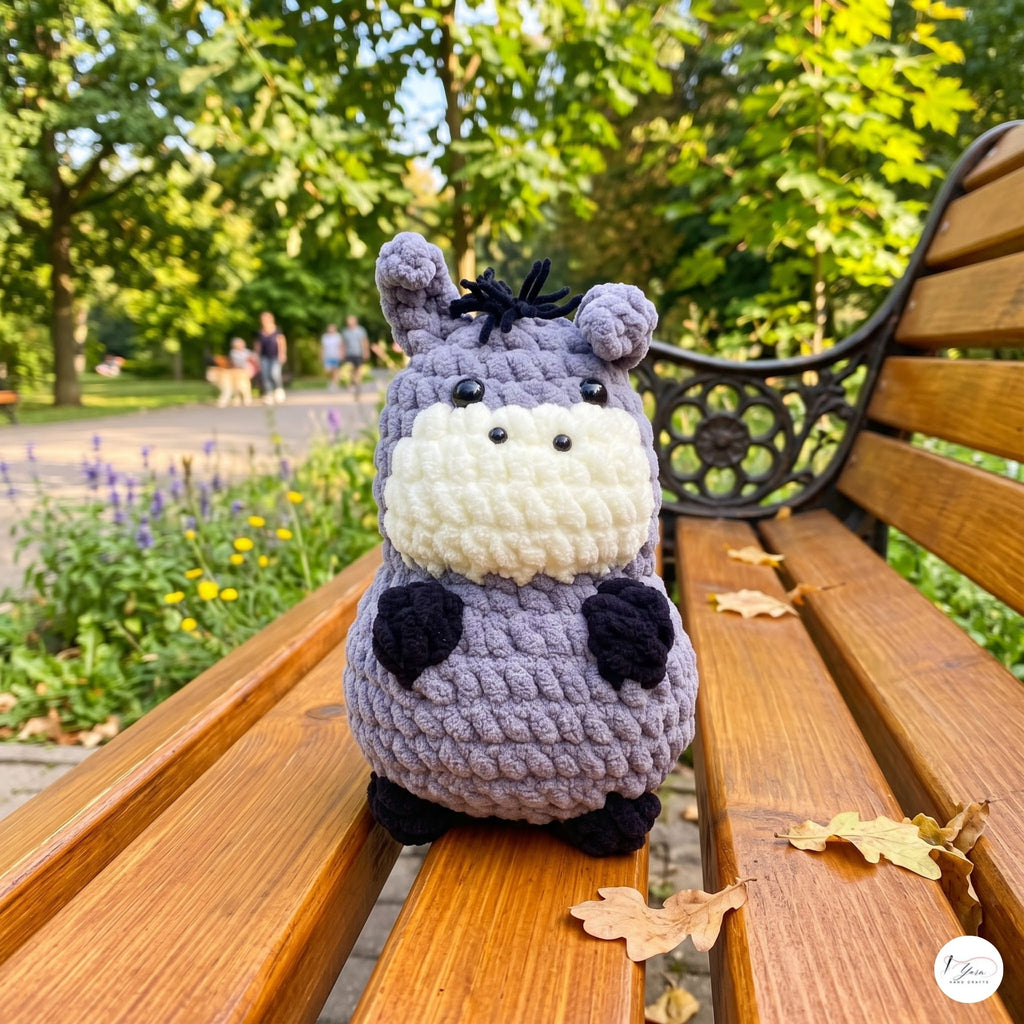 A crocheted plush toy shaped like a small donkey or hippo sits on a wooden park bench, with a blurred green garden background and a few fallen leaves nearby.