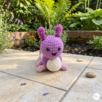 A purple and pink crocheted bunny toy with a white belly sits on a beige stone patio in a garden with green plants and flowers in the background.