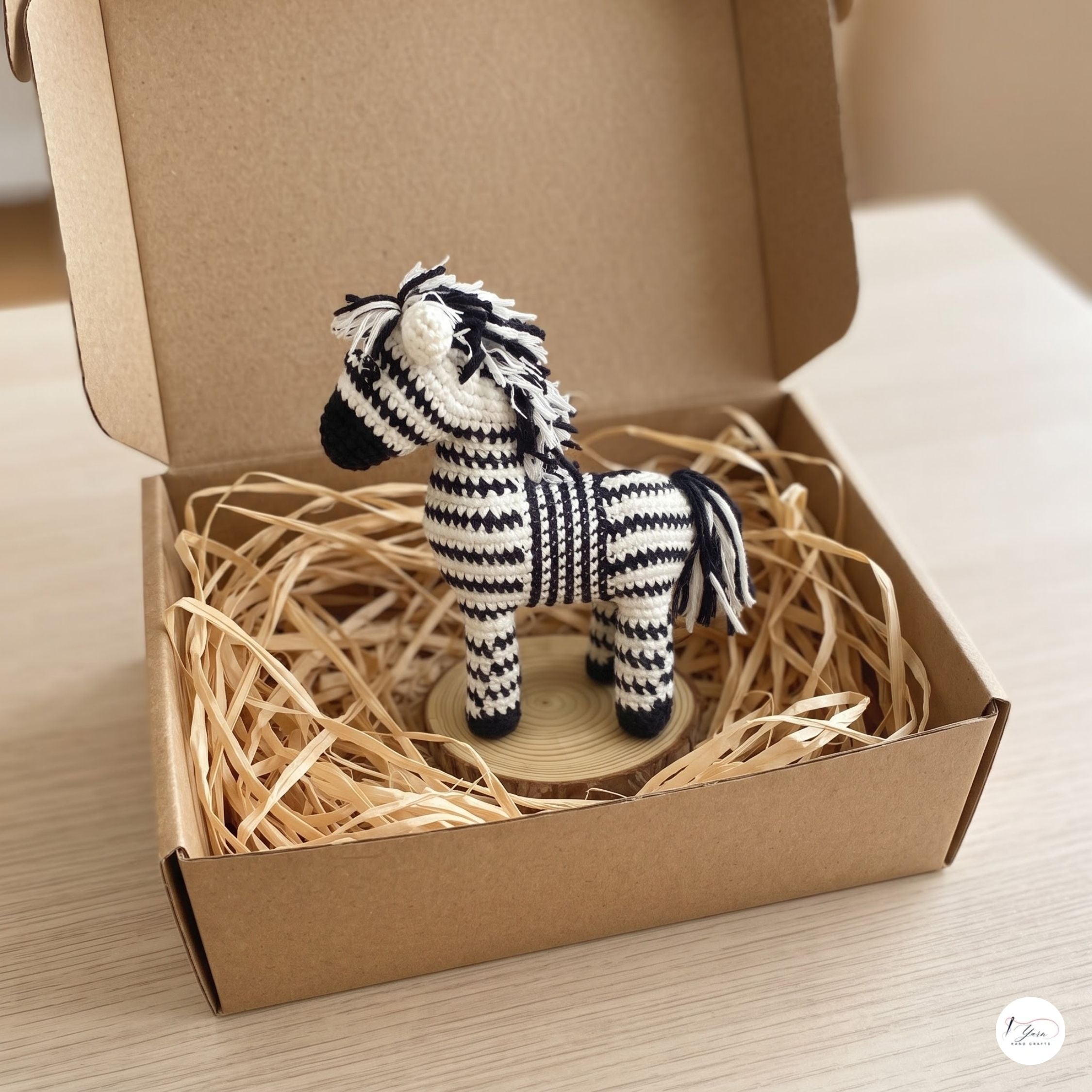 A plush handmade black and white crochet zebra toy standing on a small wooden slice inside an open cardboard gift box filled with natural brown shredded paper.